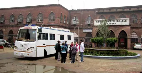 Mobile dental clinic bus parked in front of Christian Dental College, Ludhiana, with people gathered nearby.