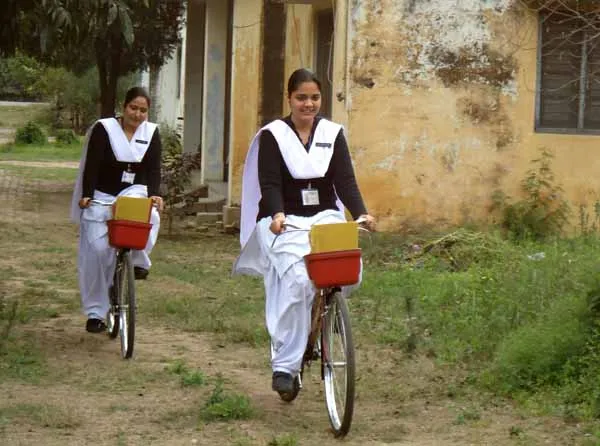 Two nursing students in white uniforms riding bicycles with medical kits for rural home health visits.
