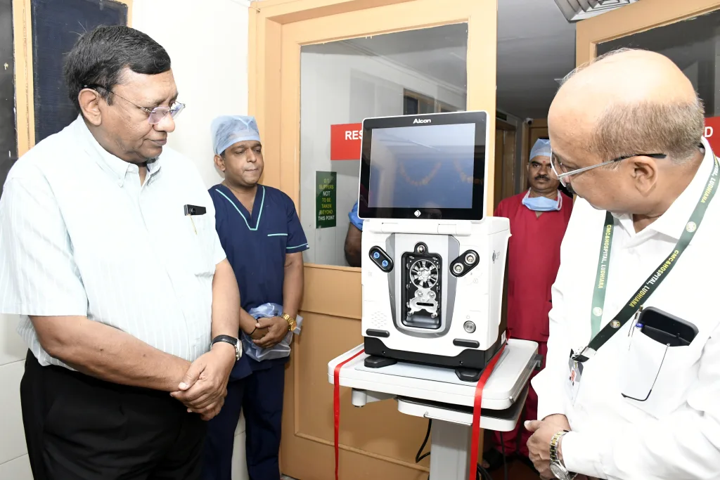 Three men stand in a hospital room, observing a medical device with a screen on a cart. One man wears medical scrubs and a cap, while the others are in formal attire. A red ribbon is tied to the cart.