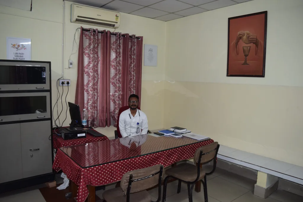 A man in a white shirt sits behind a glass-top desk with a red polka-dot tablecloth in an office. There are three chairs in front of the desk, a computer, shelves, and a painting on the wall.