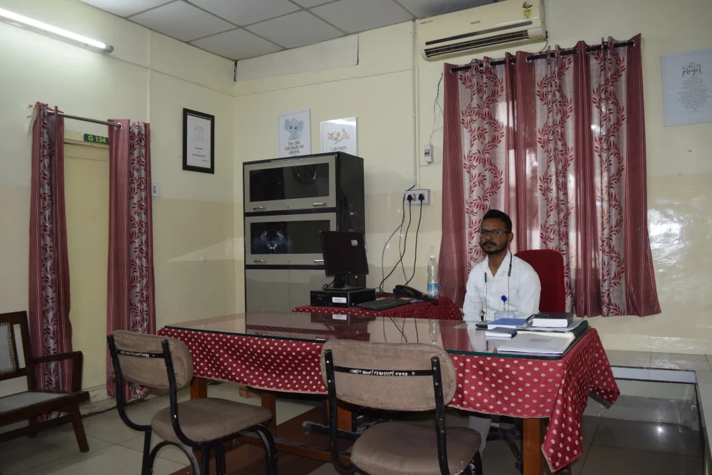 A man wearing glasses and a white coat sits at a desk with a computer and files in a small, tidy office with red curtains, a window, chairs, and a cabinet.