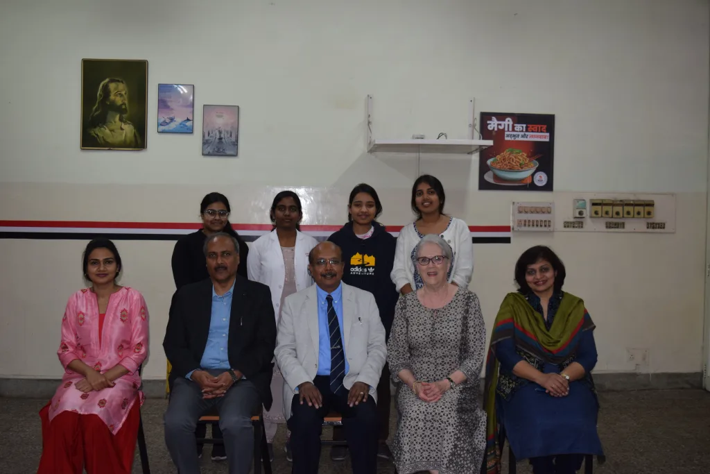 A group of nine people, including men and women, sit and stand in two rows indoors. They are dressed in formal and semi-formal attire, smiling at the camera. Pictures and a shelf are visible on the wall behind them.