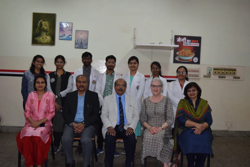 A group of eleven people, including students in white coats and adults in formal or traditional attire, pose indoors in two rows. Framed pictures and a food poster are hung on the wall behind them.