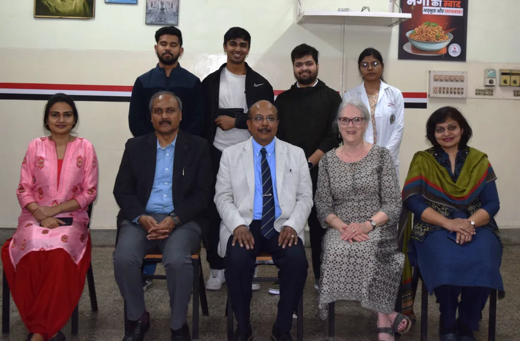 Nine people posing for a group photo indoors; four are seated in front while five stand behind them. The background includes a wall with posters, electrical switches, and a food advertisement in Hindi.
