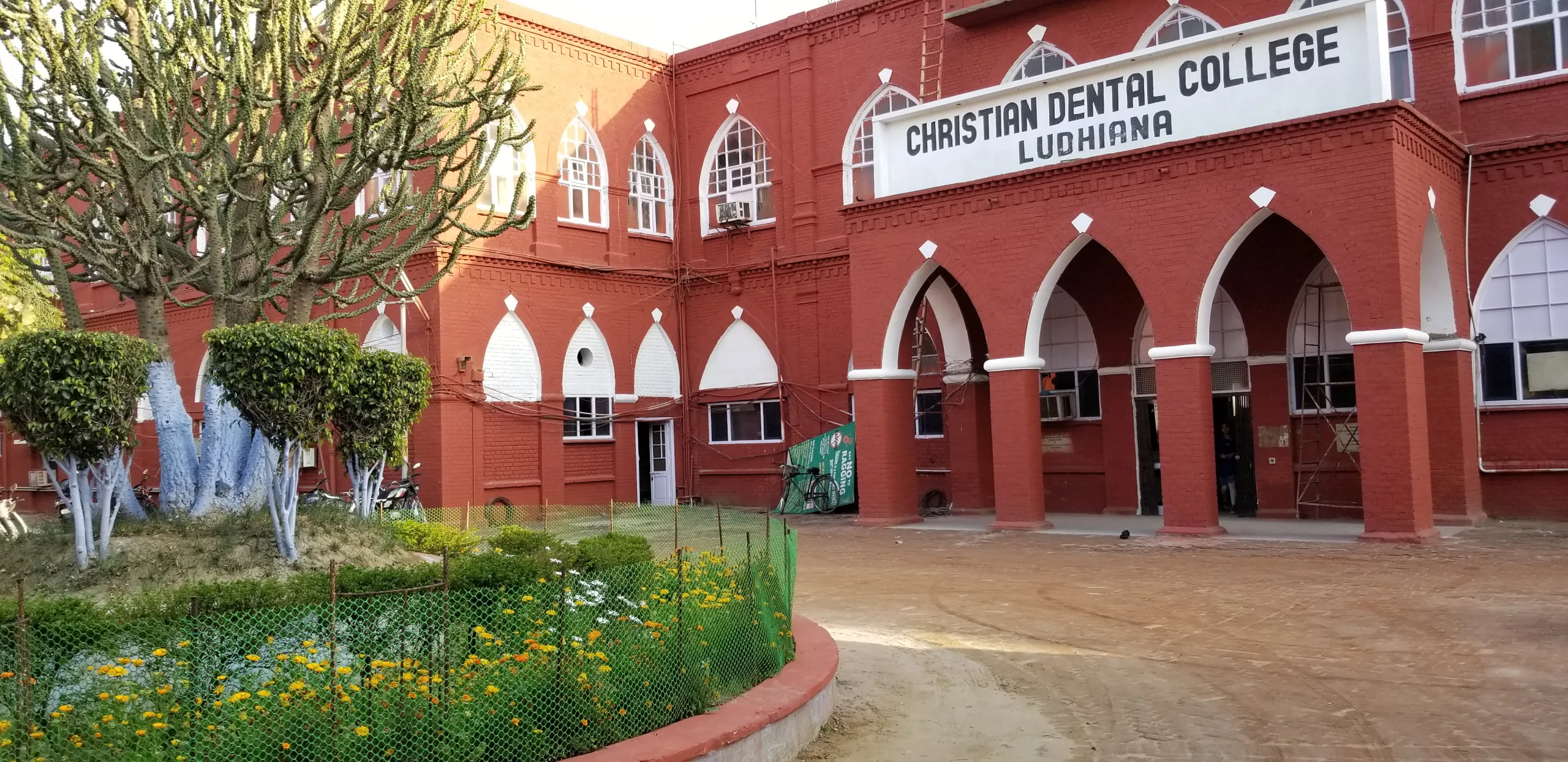 The image shows the red-brick exterior of Christian Dental College in Ludhiana, with arched doorways and windows, a sign above the entrance, and a garden with flowers and a cactus in the foreground.