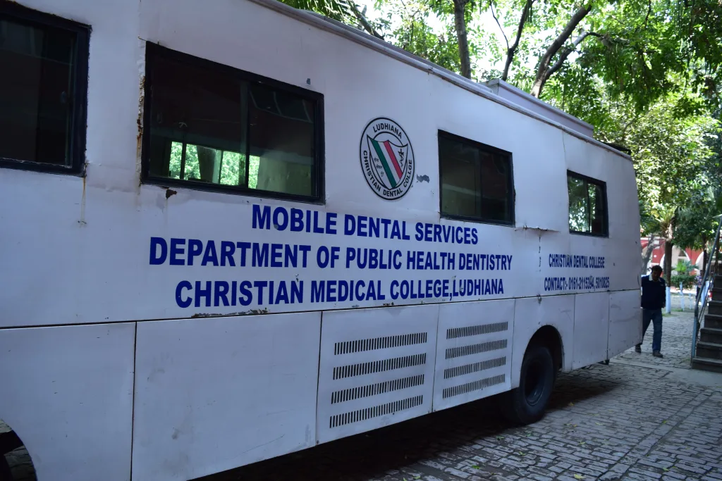 A white mobile dental services van from Christian Medical College, Ludhiana, is parked outdoors on a paved path surrounded by trees. The side of the van displays information about its dental health department.