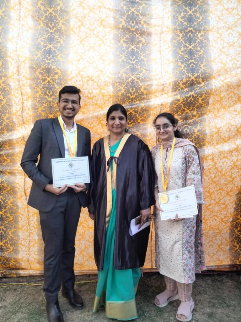 Three people stand in front of a gold patterned backdrop; the two on either side hold certificates and wear medals, while the person in the middle wears a ceremonial gown and holds papers.