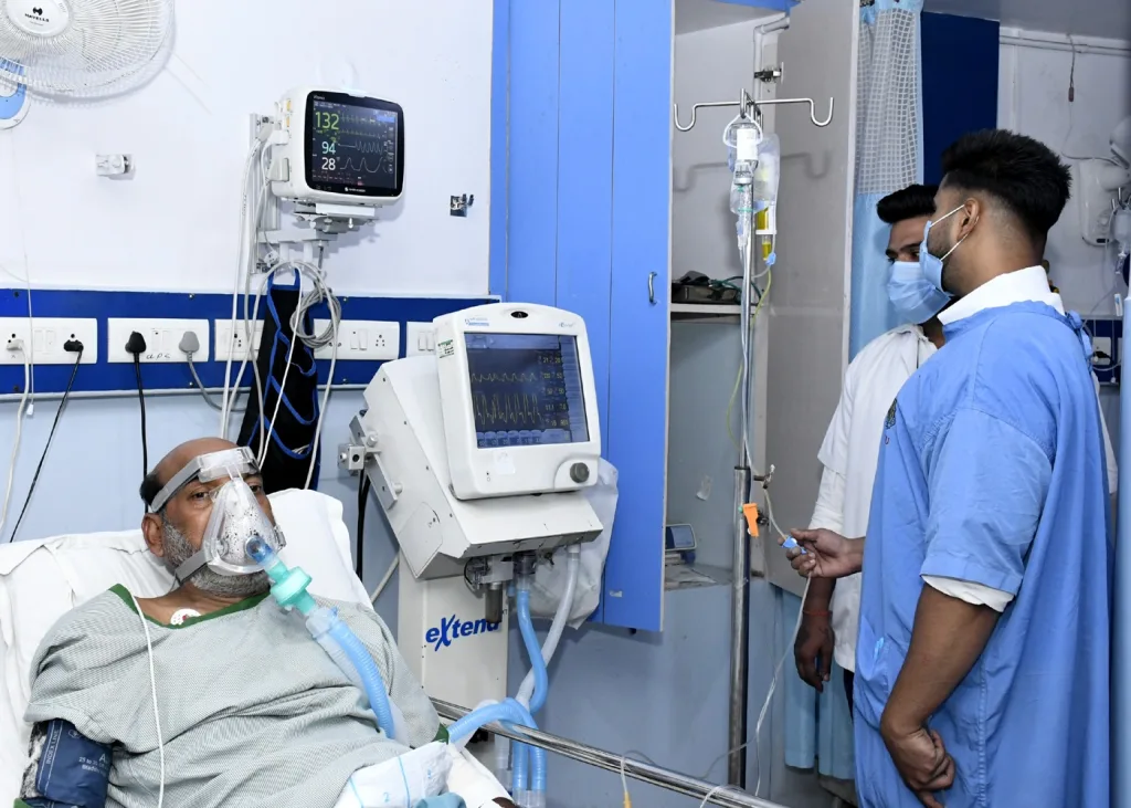 A patient wearing an oxygen mask lies in a hospital bed connected to medical equipment, while two healthcare workers in masks and gowns stand nearby monitoring his condition.