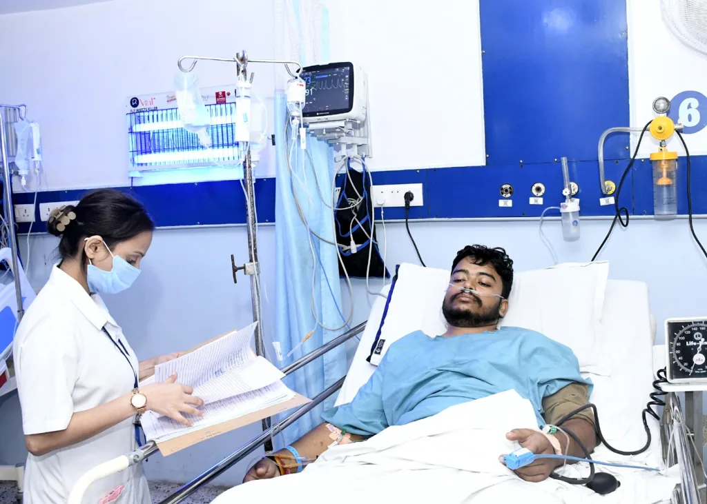 A nurse wearing a mask checks a patients chart beside a man lying in a hospital bed with monitors, IV lines, and medical equipment in a hospital room.