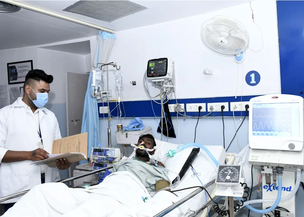 A patient lies in a hospital bed with oxygen support and monitoring equipment, while a healthcare worker in a mask reviews medical notes beside him in a clinical room.