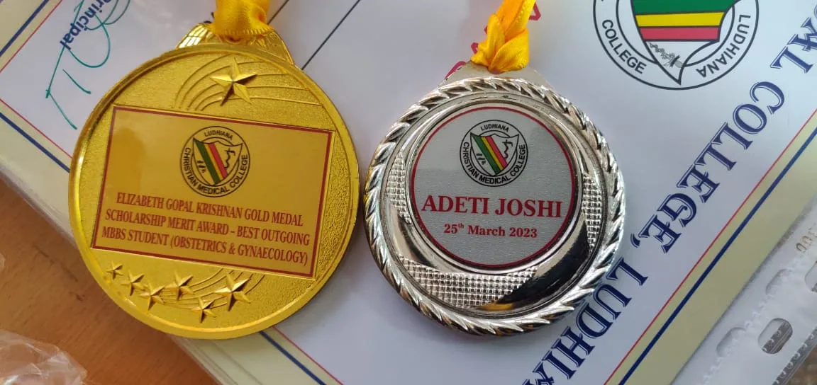 Two gold medals on yellow ribbons. The left medal reads: ELIZABETH GOPAL KRISHNAN GOLD MEDAL SCHOLARSHIP MERIT AWARD - BEST OUTGOING MBBS STUDENT (OBSTETRICS & GYNAECOLOGY). The right medal reads: ADETI JOSHI 25th March 2023.