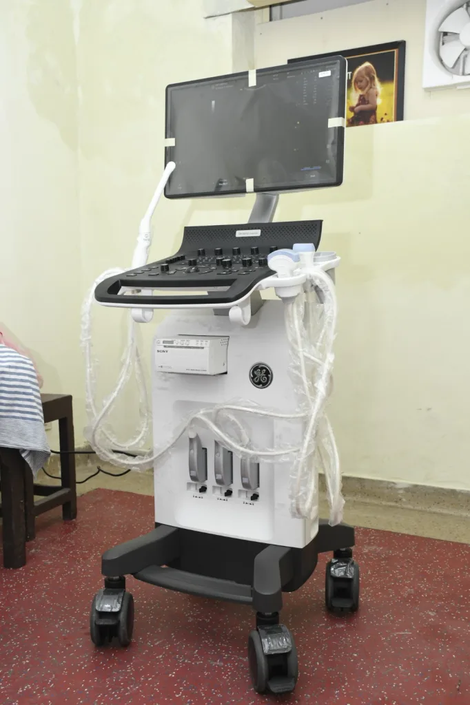 An ultrasound machine with a large monitor and multiple probes, wrapped in plastic, is positioned on a red-speckled floor in a room with light yellow walls. Framed photos hang on the wall in the background.