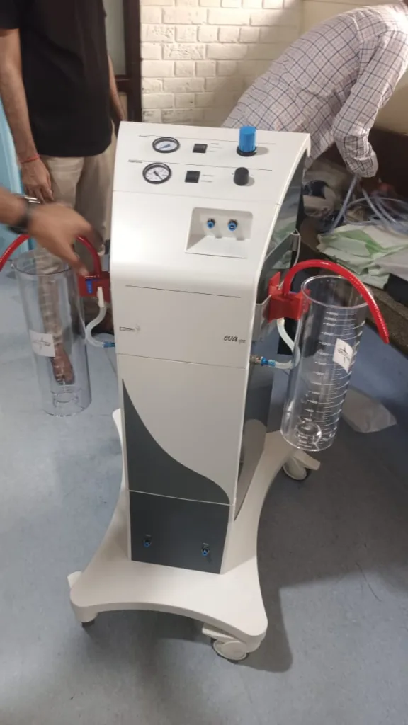A medical suction machine with two transparent canisters, gauges, and control knobs, positioned on a wheeled base in a clinical setting. Two people are visible in the background.