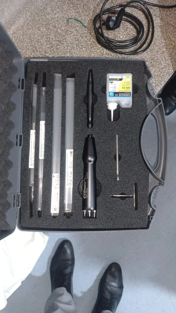 A foam-lined case containing various tools and instruments, including brushes, rods, a black handheld device, a small container of forensic ink, and other accessories. Two shoes are visible at the bottom of the image.