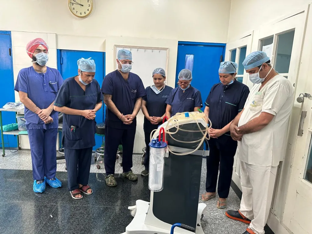 A group of six healthcare professionals in scrubs and surgical caps stand in a hospital room, heads bowed and hands folded, appearing to pray or observe a moment of silence around a medical machine.