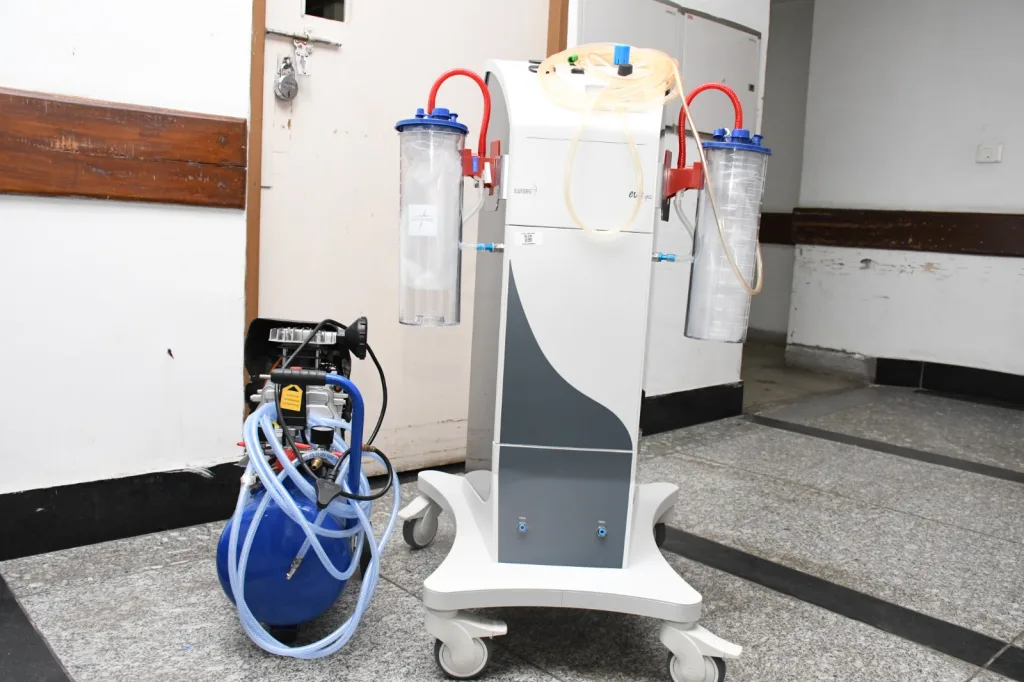 A medical suction machine with two transparent canisters and attached tubes is placed next to a blue air compressor on a wheeled base in a hospital corridor.