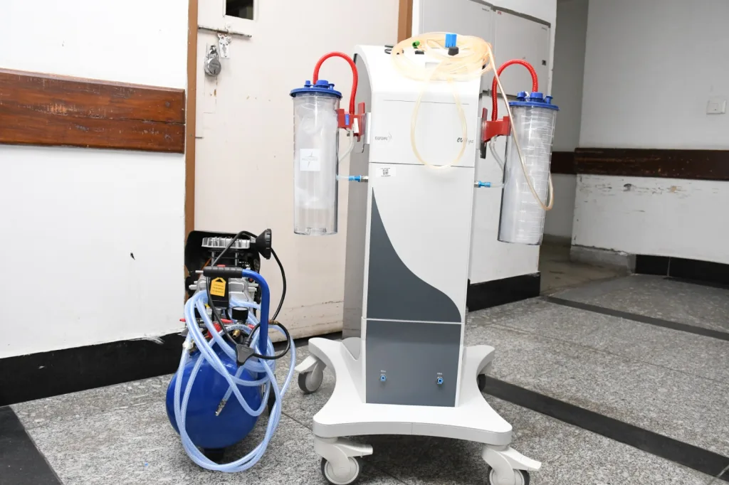 A medical suction machine with two transparent canisters, red and blue tubing, and a blue air compressor sits on a wheeled base in a hospital corridor.