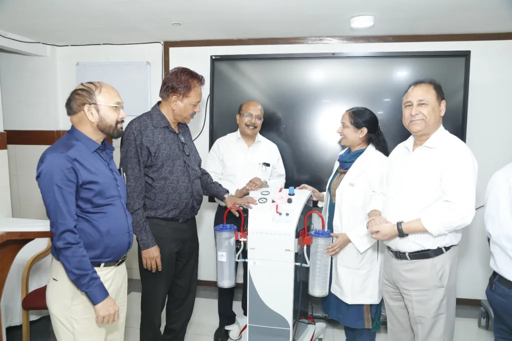 Five people, including one woman in a lab coat, stand around a medical machine with clear canisters and gauges in a well-lit room, smiling and interacting with the device.