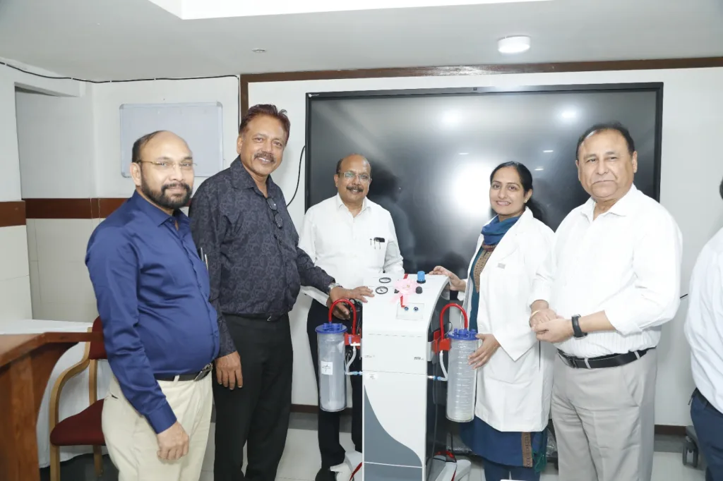 Five professionals, including two women in lab coats and three men in business attire, stand around a medical suction device in a modern room with a large monitor in the background. They are smiling and posing for the photo.
