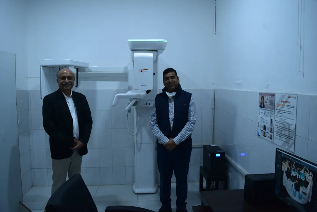 Two men stand in a tiled room beside a dental X-ray machine. One man wears a suit, the other a checkered shirt with a vest. A desk with a computer and posters on the wall are also visible.