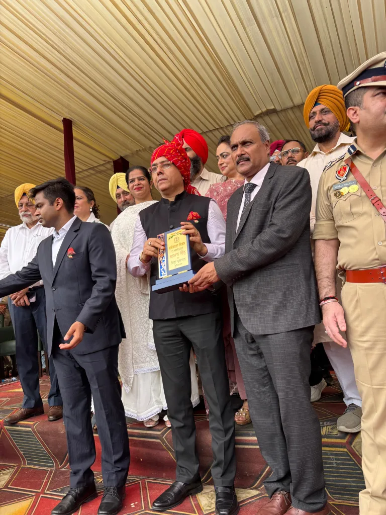 A group of people, including men in suits and uniforms, stand together at an event at Christian Medical College in Ludhiana. One man in a red turban holds a plaque, surrounded by others on a patterned floor under a covered area.