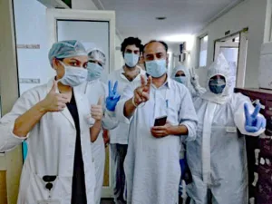 A group of healthcare workers at Christian Medical College in Ludhiana, wearing masks and protective gear, stand in a hospital corridor making peace and thumbs-up signs while looking at the camera.