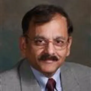 A man with short dark hair, a mustache, and glasses, wearing a gray suit jacket, white shirt, and maroon tie, poses in front of a plain dark background, presenting a professional look suitable for the Board of Directors.