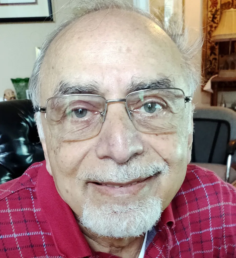 An elderly man with glasses, a white beard, and a mustache smiles at the camera. Wearing a red plaid shirt, he sits indoors in a well-lit room, exuding the warmth and wisdom often found among Board of Directors members.