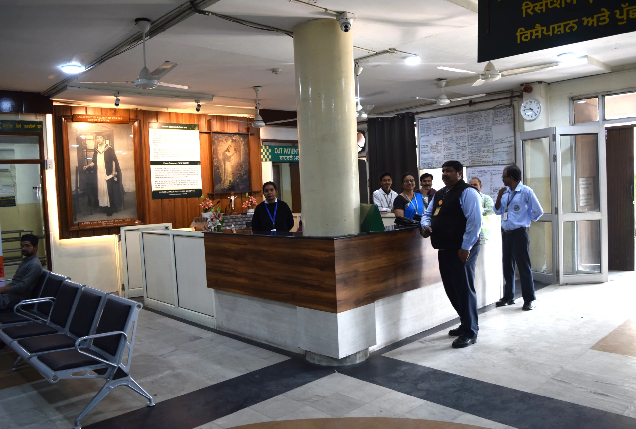 A hospital reception area at Christian Medical College in Ludhiana, with uniformed staff behind the counter, a security guard in front, and empty chairs neatly arranged in the waiting area.