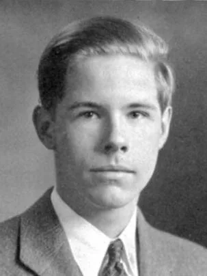 Black and white portrait of a young man with short, neatly combed hair, wearing a suit jacket, dress shirt, and patterned tie, looking directly at the camera with a neutral expression.