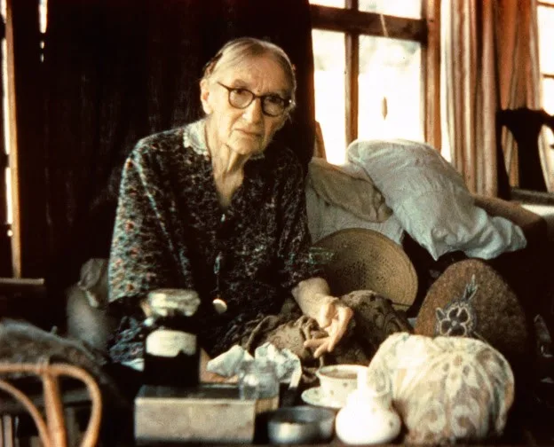 An elderly woman with glasses and gray hair sits indoors on a cluttered couch, surrounded by pillows, jars, and teacups, with a window and curtains in the background.