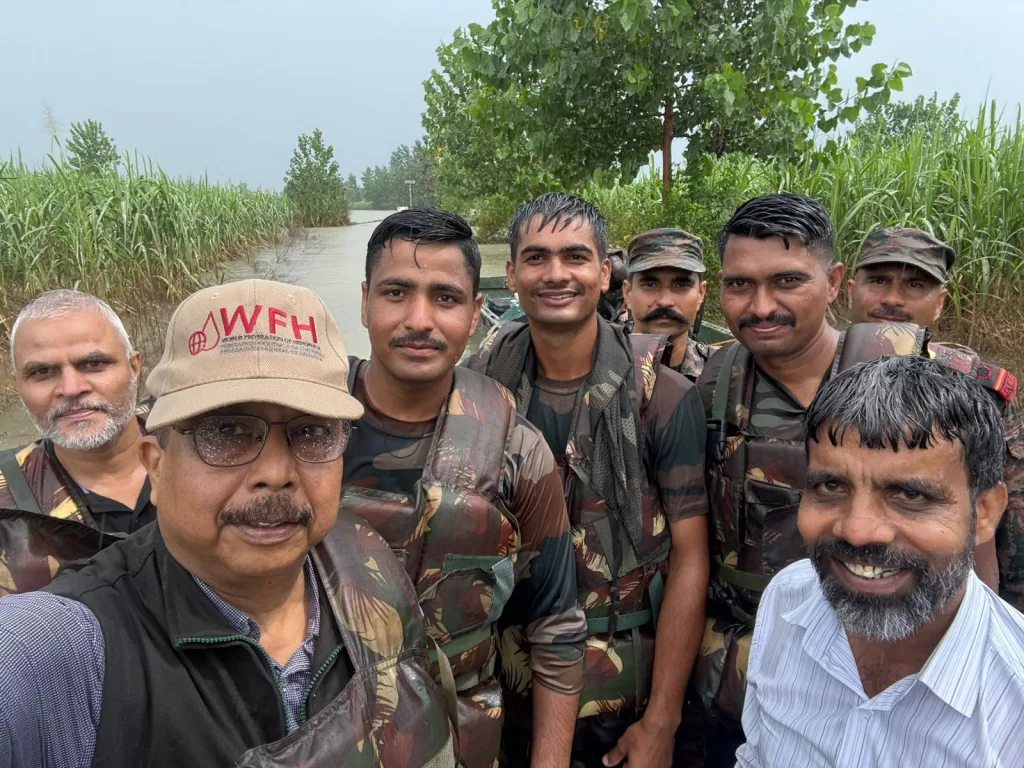 A group of men, some in camouflage uniforms and life jackets, stand together outdoors near water and greenery, smiling at the camera. One man in front wears glasses and a tan cap with red WFH text.
