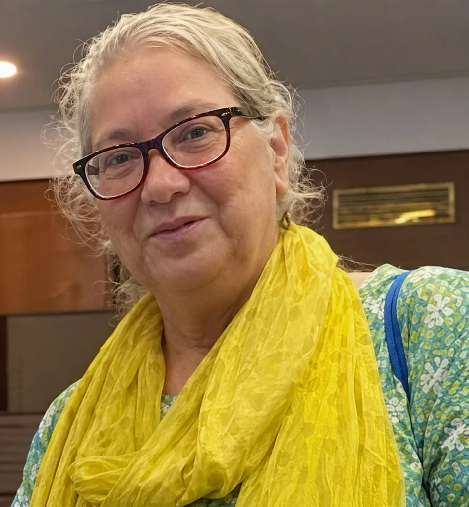 An older woman with gray hair, glasses, and a yellow scarf over a green patterned top smiles at the camera indoors, reflecting the warmth and professionalism often seen among Board of Directors members.
