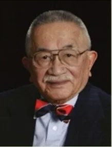 An elderly man with short gray hair, glasses, and a mustache, wearing a dark suit, white shirt, and a red and blue bow tie, poses against a dark background.