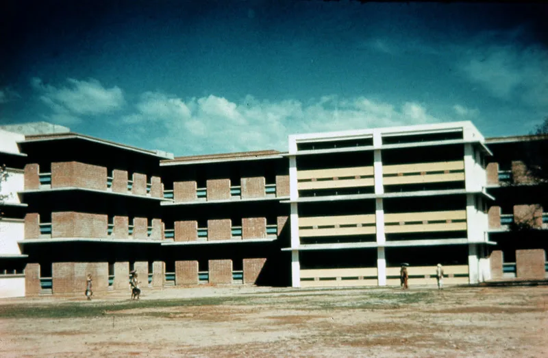 A modern three-story brick and concrete building with horizontal windows, set against a blue sky with scattered clouds. A few people walk across the open ground in front of the structure.