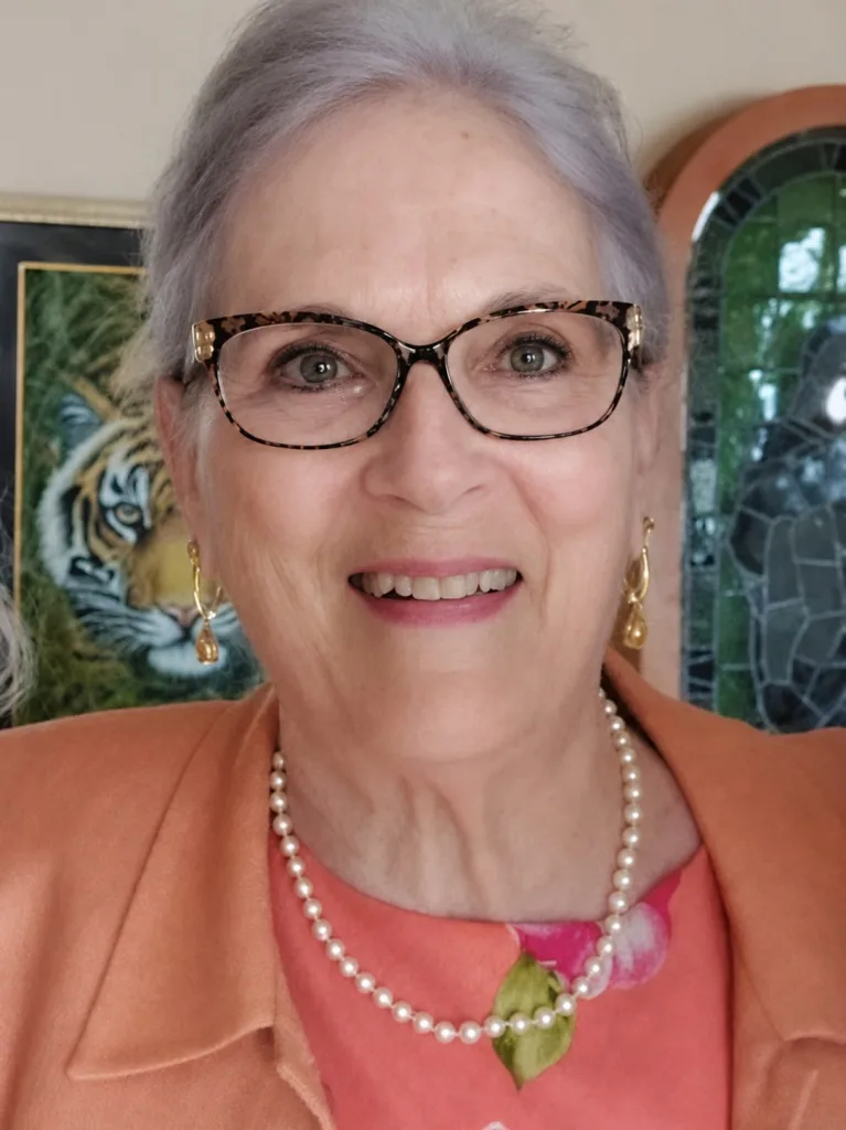 An older woman with gray hair, wearing glasses, gold earrings, a pearl necklace, and an orange jacket over a floral top, smiles at the camera. A Board of Directors plaque, a tiger painting, and mosaic artwork are visible in the background.