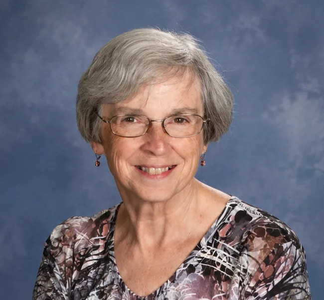 An older woman with short gray hair and glasses smiles at the camera. She is wearing a patterned top and earrings, with a soft blue studio backdrop behind her.